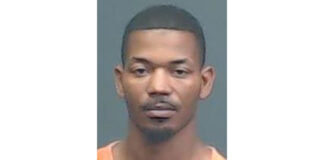 A man with short hair and a trimmed goatee wears an orange shirt and looks straight ahead against a plain, dark background.