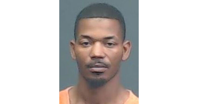 A man with short hair and a trimmed goatee wears an orange shirt and looks straight ahead against a plain, dark background.
