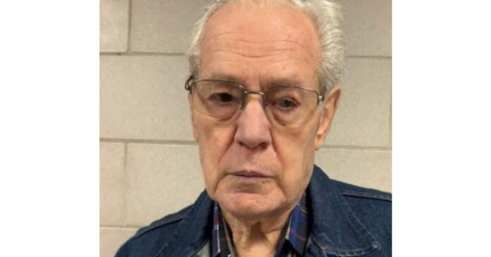 Mugshot of An older man with gray hair and glasses wearing a blue denim jacket and a plaid shirt, standing in front of a light-colored brick wall.