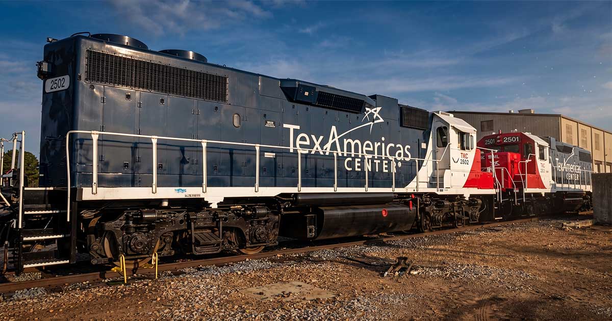 A side-view photograph of a dark blue diesel locomotive, engine number 2502, featuring a prominent white "TexAmericas CENTER" logo on its side. It is coupled to a red and white locomotive, engine number 2501, sitting on gravel-lined tracks on a sunny day.