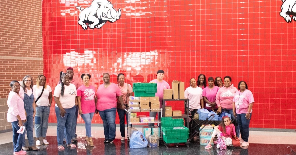 Arkansas High School Class of 1991 members in pink shirts stand with carts of donated food in front of a red Razorback wall