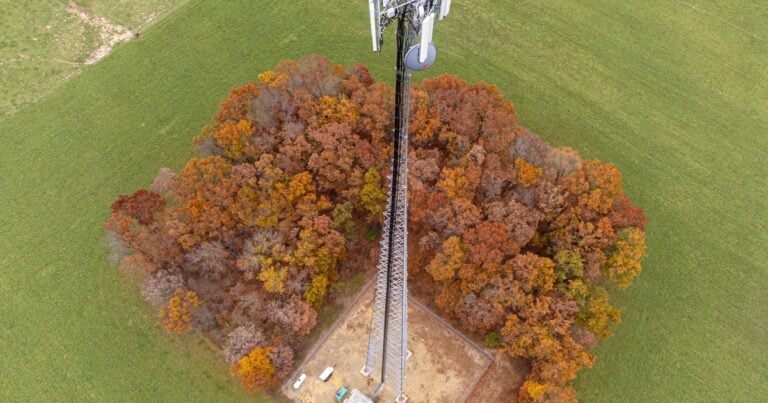 Aerial view of a lattice cell tower inside a fenced compound surrounded by trees