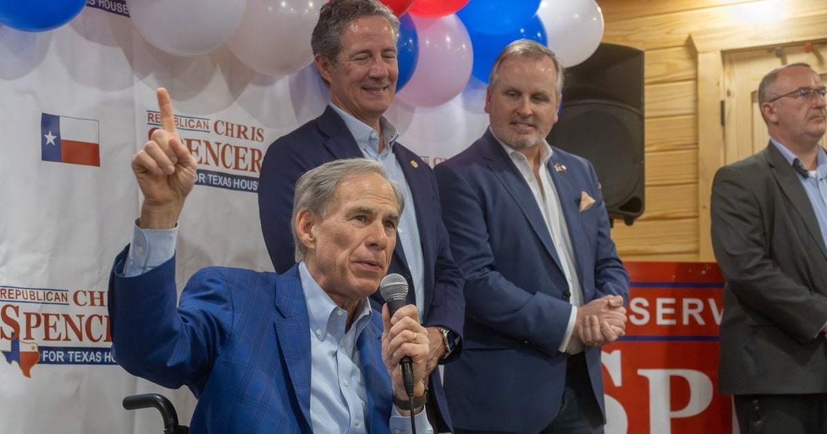 Gov. Greg Abbott, seated with a microphone and raising one finger, speaks in front of Chris Spencer for Texas House campaign signs and red, white and blue balloons