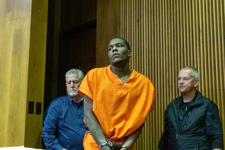 Omarion Owens in an orange jail uniform and handcuffs, escorted by two men in a Bowie County courtroom