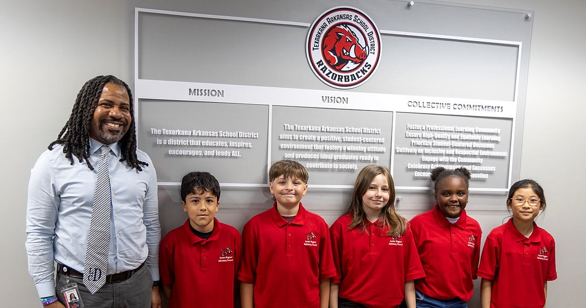 Larry Dunn stands with five fifth graders in red Junior Magnet Advisory Council shirts in front of the TASD mission and vision wall