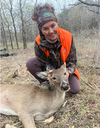 A smiling hunter in a blaze orange vest kneels beside a harvested white-tailed doe